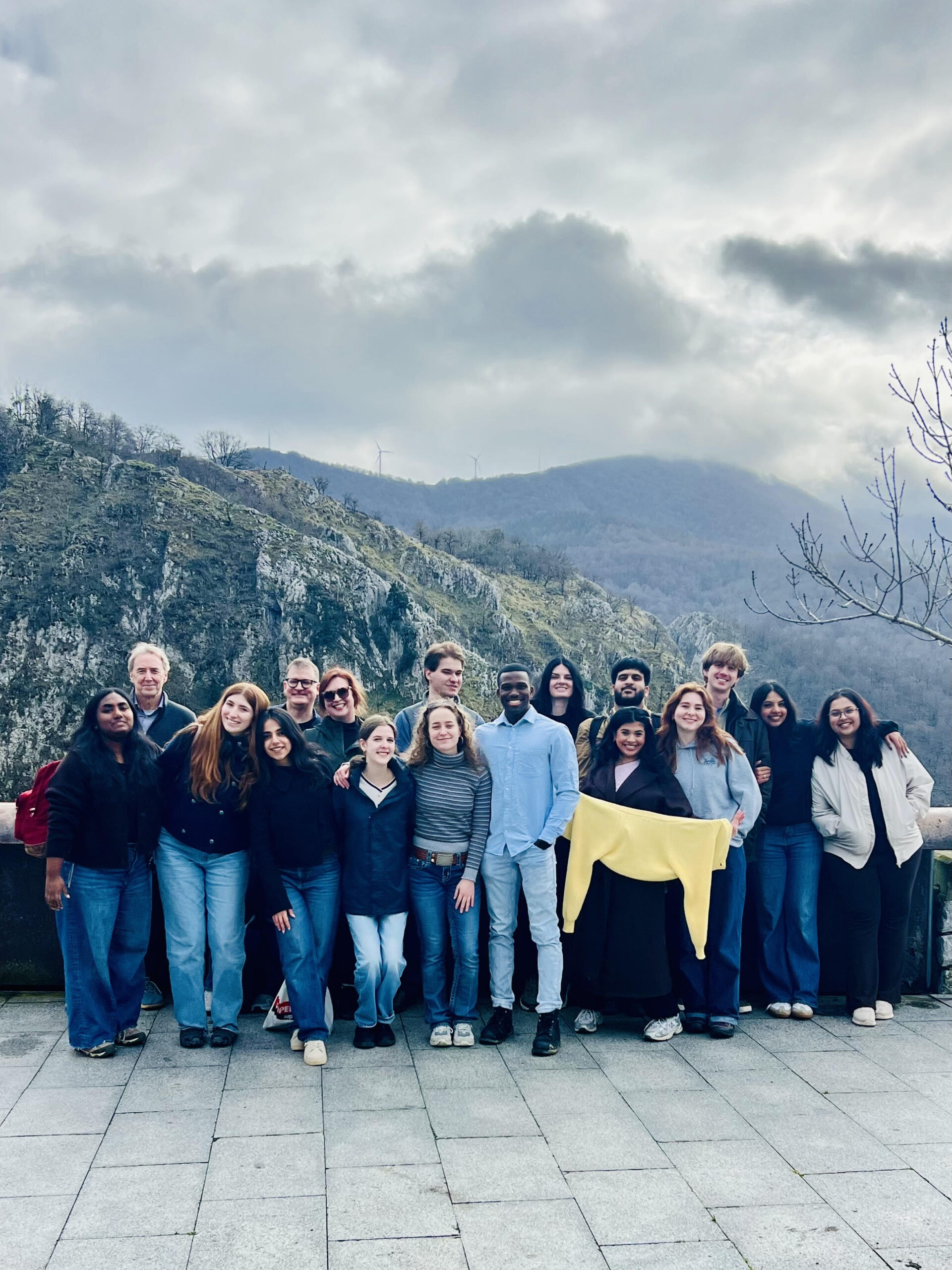 Seventeen adults, most being students, pose for a picture in front of a jutting mountain-scape in the Basque region in Spain on a cloudy day.