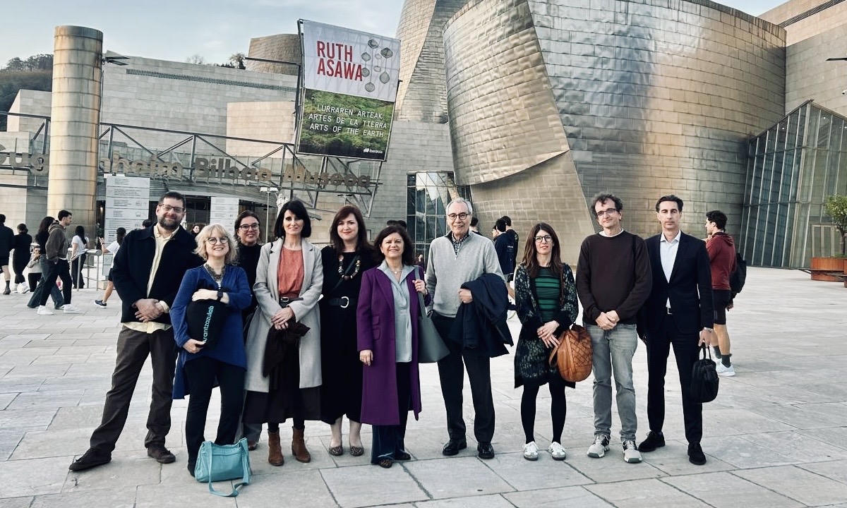 Ten professionally-dressed adults pose center-frame in front of the tinfoil-like walls of the Guggenheim Bilbao Museum.