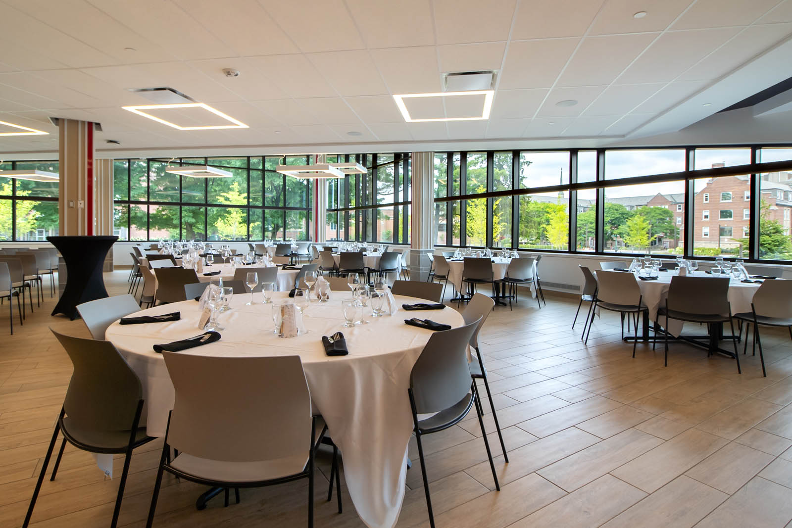 A large modern room with windows out to the quad full of green trees featuring round tables draped with tablecloths setup for a formal reception