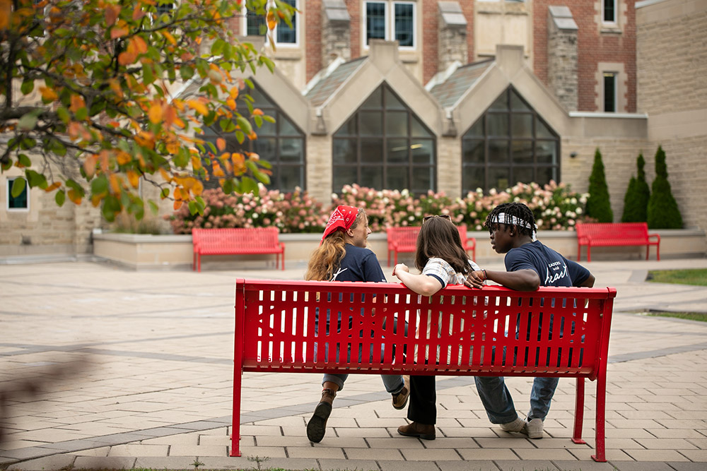 Three students sitting on a red bench on campus in the fall