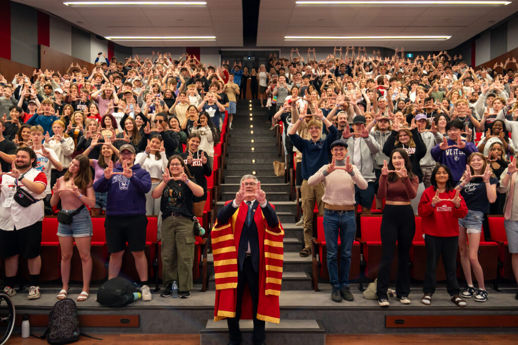 Dr Barry Craig standing in the centre of the Huron Theatre making the H sign with his fingers with the incoming 2024 class making the same sign in the background