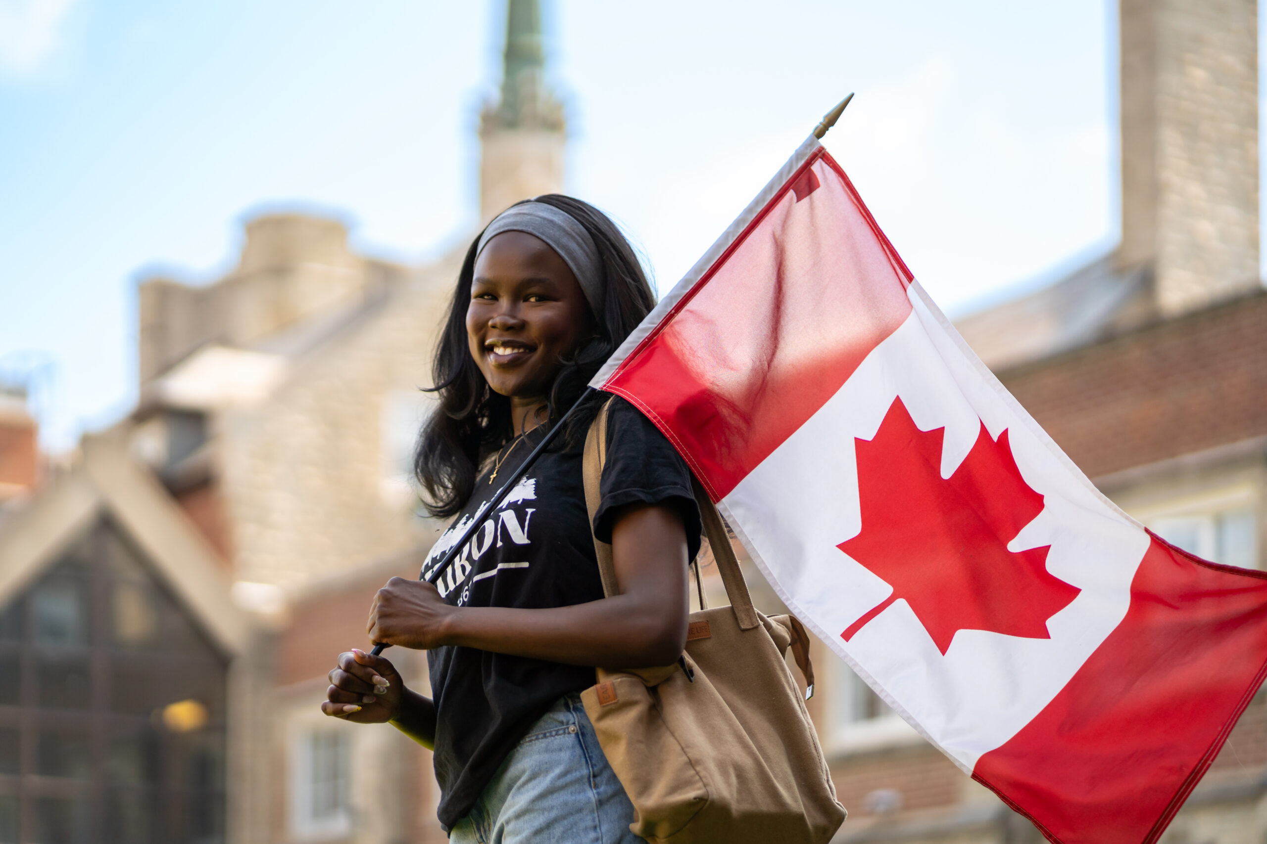 A dark-skinned woman smiles over her shoulder as a Canadian flag flaps from the pole that she's holding. Huron's chapel can be seen in the background.